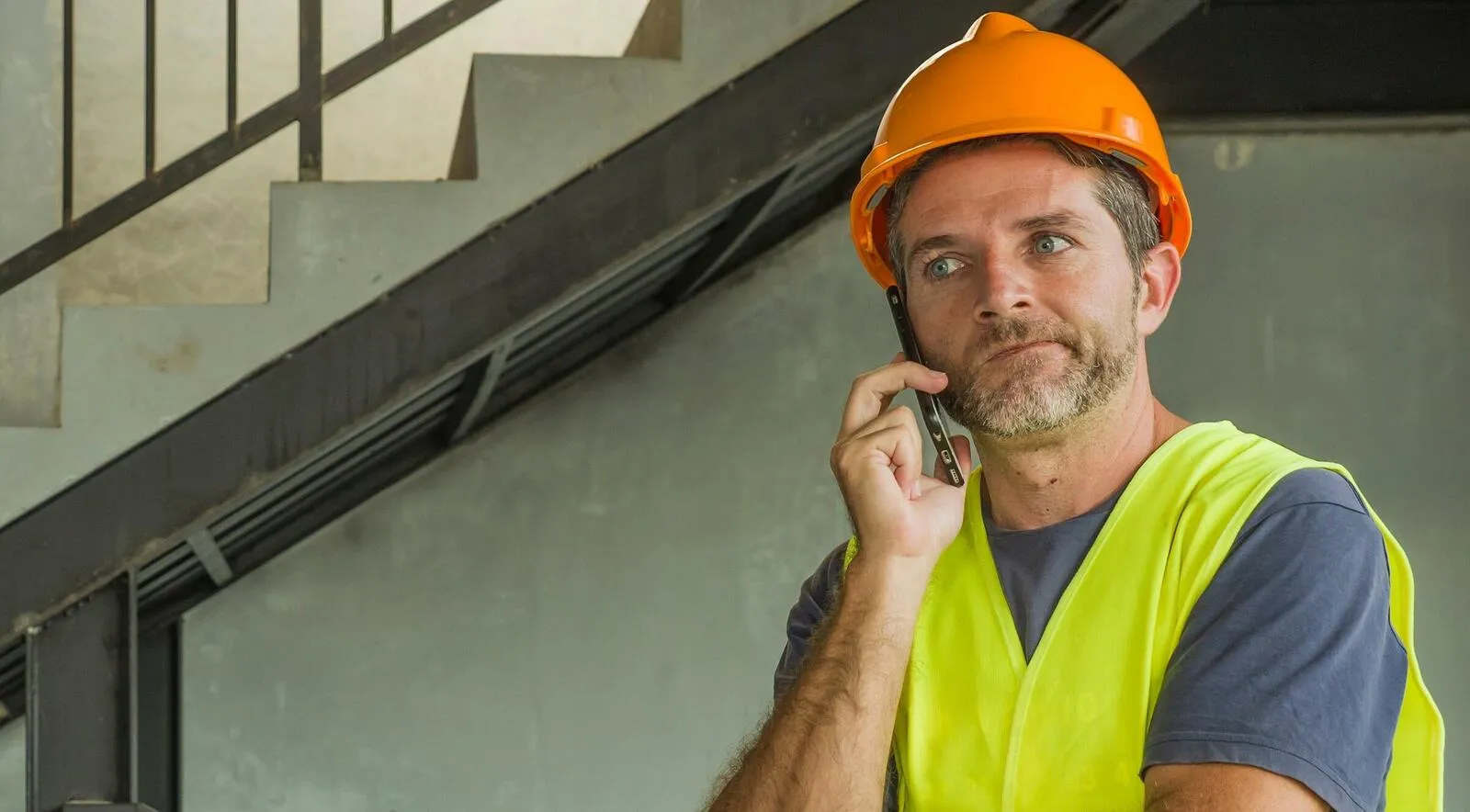 Ein Handwerker mi einem Schutzhelm und einer gelben Warnweste hält sich das Smartphone ans Ohr und guckt nachdenklich.